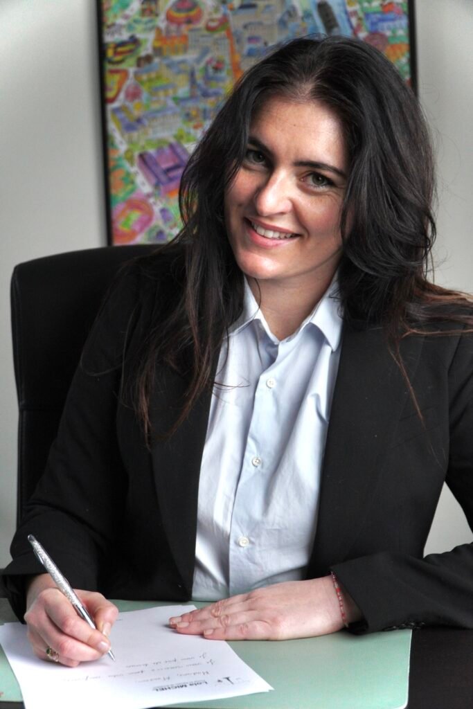 Photographie d'une jeune femme brune souriante, professionnelle, assise à un bureau élégant. Elle porte une chemise blanche et un blazer noir, tenant un stylo, prête à signer ou rédiger un contrat juridique, symbolisant un environnement de travail sérieux et formel.